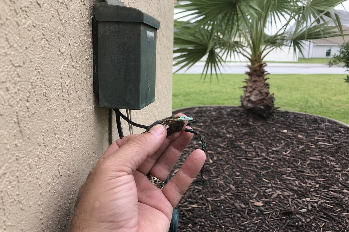 Close-up of a corroded low-voltage wire connection being held next to a green landscape lighting transformer mounted on a Florida stucco wall